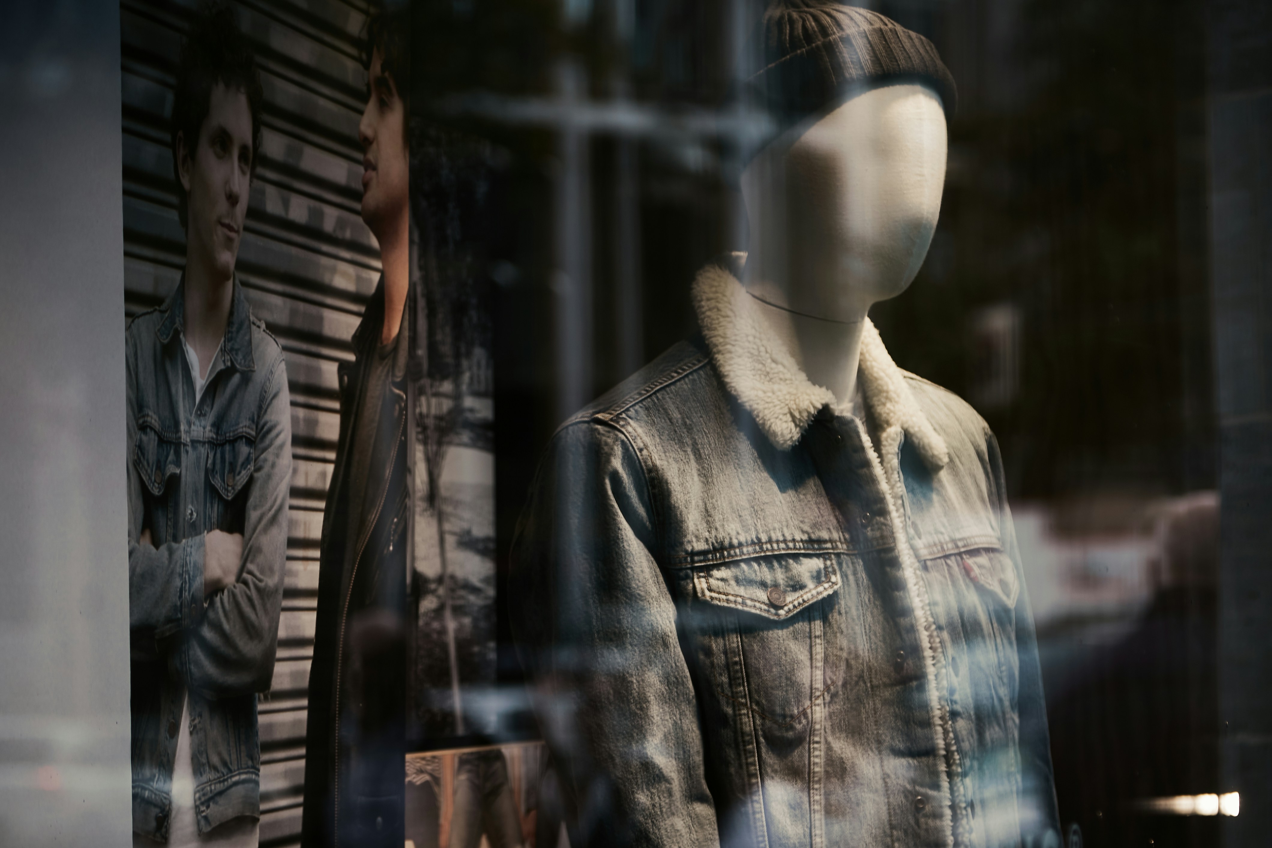 Mannequin wearing a denim jacket with shearling collar and beanie in a fashion store window display, with reflections and urban streetwear posters in the background.