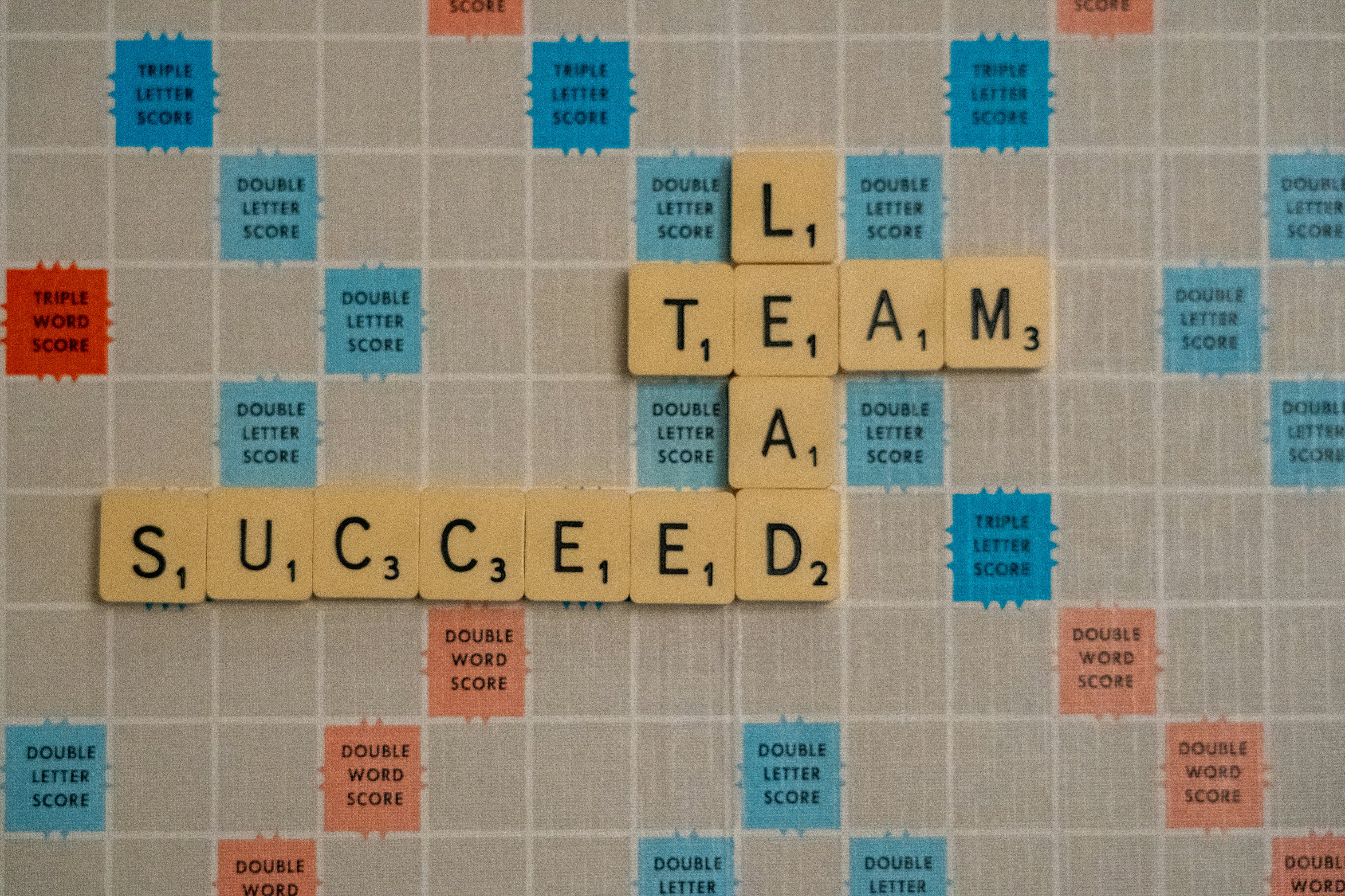 Scrabble game board with letter tiles forming the words “LEAD,” “TEAM,” and “SUCCEED,” symbolizing teamwork and leadership as a path to success.